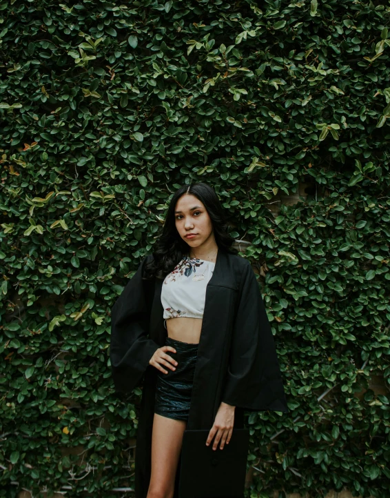 a woman standing in front of a green wall, by Robbie Trevino, unsplash, wearing a black robe, wearing a crop top, graduation photo, clad in vines