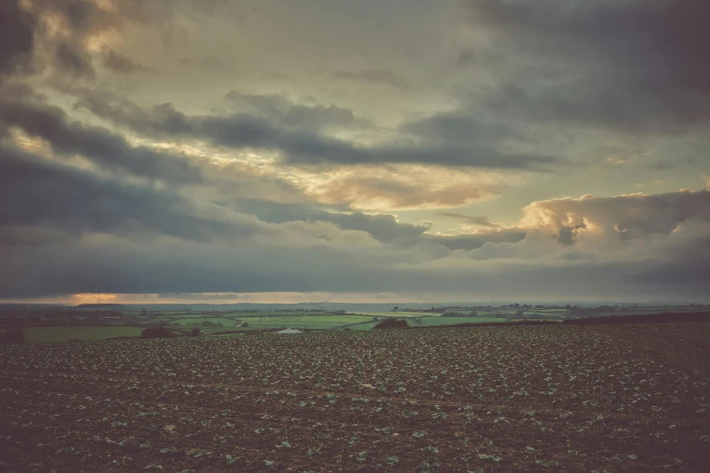 a cloudy sky over a plowed field, by Andrew Bell, unsplash, early evening, distant villagescape, in muted colours, rocky ground