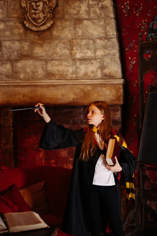 a woman standing in a living room holding a wand, inspired by Hermione Hammond, pexels contest winner, academic art, wearing an academic gown, in an action pose, school class, in front of a fireplace