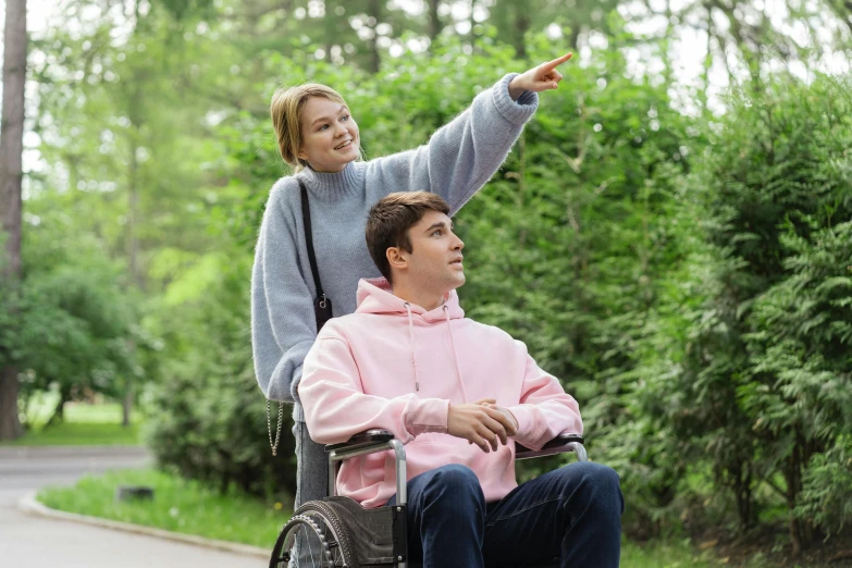 a woman pointing at a man in a wheelchair, trending on pexels, hurufiyya, wearing a pastel pink hoodie, nature outside, gray, nursing