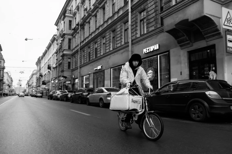 a woman riding a bike down a city street, a black and white photo, by Emma Andijewska, pexels contest winner, realism, getting groceries, saint petersburg, action shot girl in parka, delivering parsel box