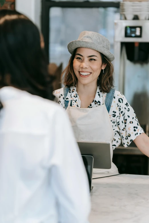 a woman standing behind a counter at a restaurant, trending on pexels, happening, smiling at each other, aussie baristas, woman with hat, asian female