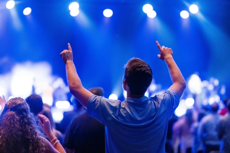 a man standing in front of a crowd with his hands in the air, blue light accents, listening to godly music, college, looking from shoulder