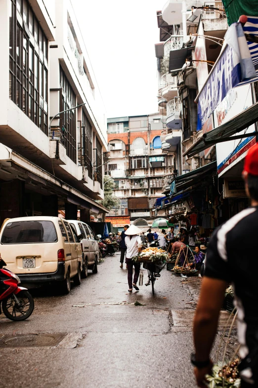 a man riding a bike down a street next to tall buildings, wet market street, travelers walking the streets, old buildings, full of trash