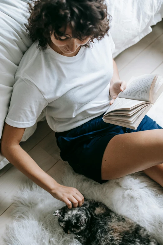 a woman sitting on a bed reading a book next to a dog, trending on unsplash, renaissance, tan skin a tee shirt and shorts, white fluffy cotton shorts, lying down, holding books