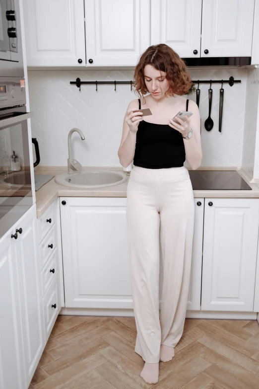 a woman standing in a kitchen looking at her cell phone, by Grytė Pintukaitė, unsplash, renaissance, large pants, pale milky white porcelain skin, pale fair skin!!, 155 cm tall