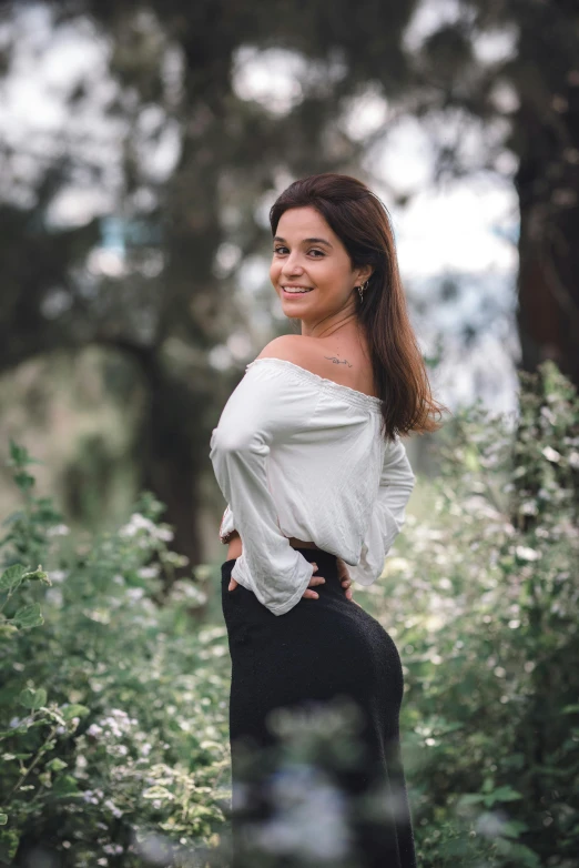 a woman standing in the middle of a forest, by Charly Amani, wearing tight shirt, elegant smiling pose, around 20 yo, showing her shoulder from back