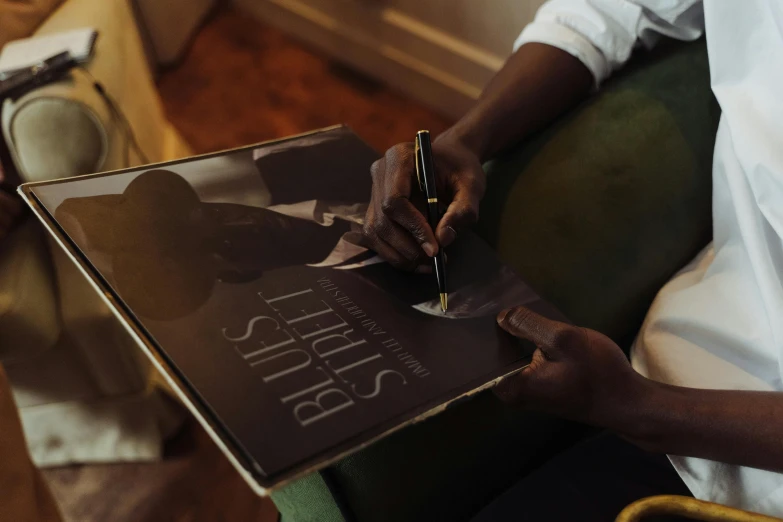 a person sitting in a chair writing on a book, an album cover, inspired by Gordon Parks, pexels contest winner, black arts movement, blue steel, signatures, velvet art, a high angle shot