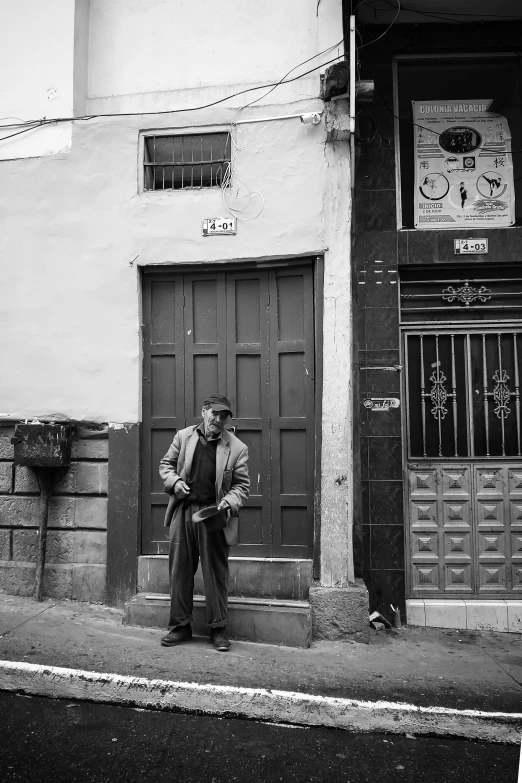 a black and white photo of a man standing in front of a building, a black and white photo, inspired by Henri Cartier-Bresson, flickr, quito school, in front of the house, old pawn shop, teruel city in 1989, holding a cane