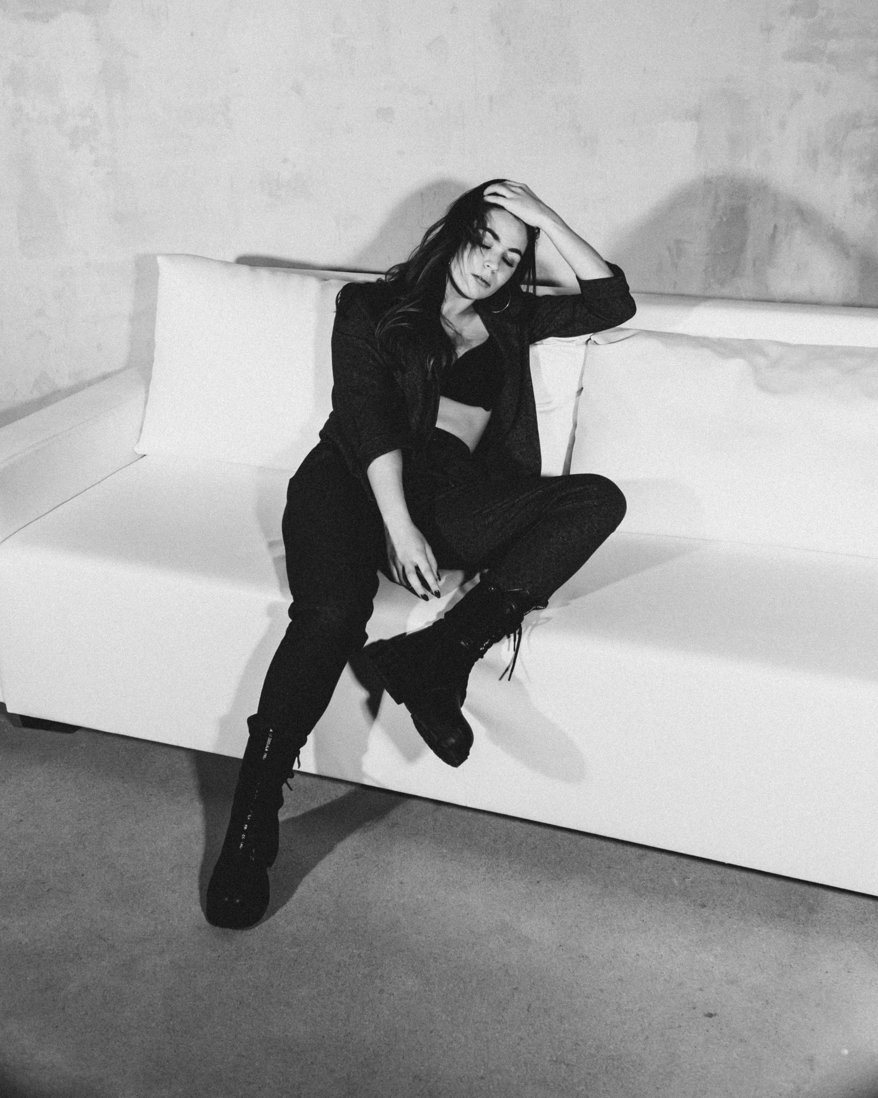 a black and white photo of a woman sitting on a couch, dua lipa, she wears boots, leaning against the wall, olga buzova