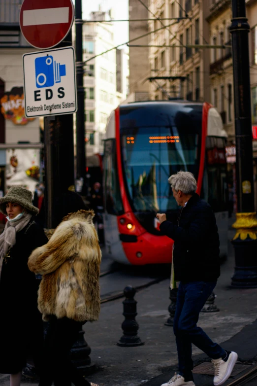 a group of people crossing a street in a city, a picture, by Raphaël Collin, street tram, wearing red fur, white haired lady, close-up photo