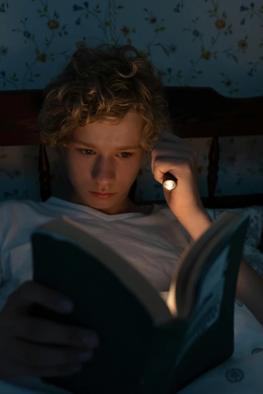 a person laying in bed reading a book, a portrait, by David Donaldson, pexels, light over boy, still from a live action movie, flashlight on, blond boy