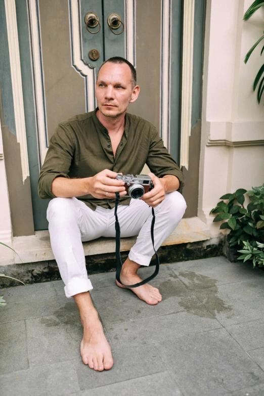 a man sitting in front of a door holding a camera, a picture, hairless, wearing a linen shirt, bali, barefoot