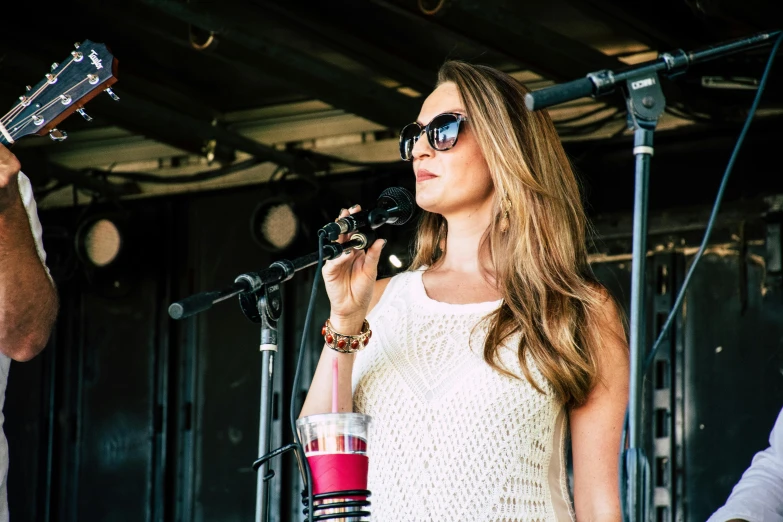 a woman playing a guitar and singing into a microphone, covered outdoor stage, drink, looking off to the side, olivia