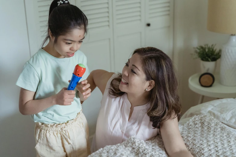 a woman sitting on top of a bed next to a little girl, inspired by Ruth Jên, pexels contest winner, happening, singing into microphone, fisher price, holding maracas, asian female