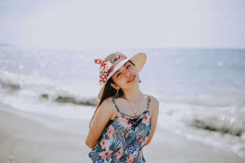 a woman standing on top of a beach next to the ocean, with cute - fine - face, floral clothes, it's wearing a cute little hat, south east asian with round face