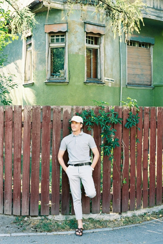 a man leaning against a fence in front of a house, an album cover, inspired by Bencho Obreshkov, pexels contest winner, wearing polo shirt, full body photo, wearing a baseball cap, slavic