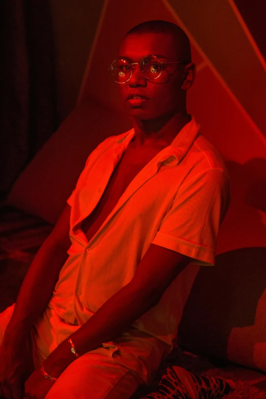 a man sitting on top of a bed under a red light, pexels contest winner, afrofuturism, man with glasses, non binary model, ( ( dark skin ) ), lean man with light tan skin