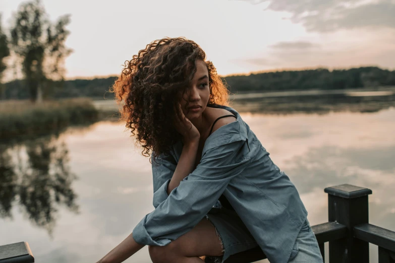 a woman sitting on a dock next to a body of water, trending on pexels, happening, brown curly hair, portrait sophie mudd, mixed-race woman, during dawn