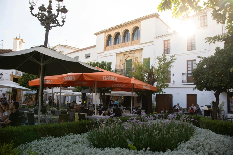 a group of people sitting under umbrellas in a courtyard, marbella, white marble buildings, white and orange, restaurant in background