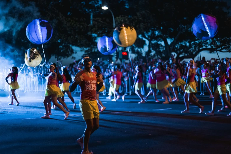 a group of people walking down a street at night, orange and yellow costume, sydney park, blue and red lights, float