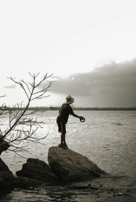 a man standing on top of a rock next to a body of water, by Sunil Das, pexels contest winner, windy mood, aboriginal capirote, “ iron bark, teenage boy