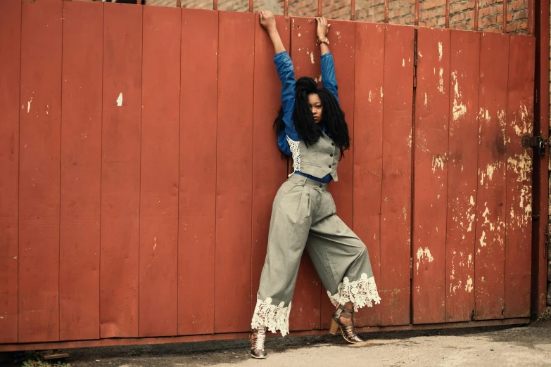 a woman leaning against a fence with her arms in the air, an album cover, by Lily Delissa Joseph, trending on pexels, arabesque, artist wearing overalls, wide leg hakama trousers, brown skinned, grey