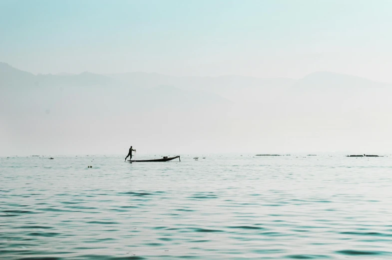 a man riding a paddle board on top of a body of water, a picture, unsplash contest winner, minimalism, fishing boats, myanmar, muted color (blues, hazy water