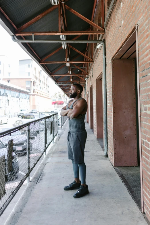 a man standing on a sidewalk next to a building, by Andrew Stevovich, wearing fitness gear, jaylen brown, ponytail and beard, looking out the window