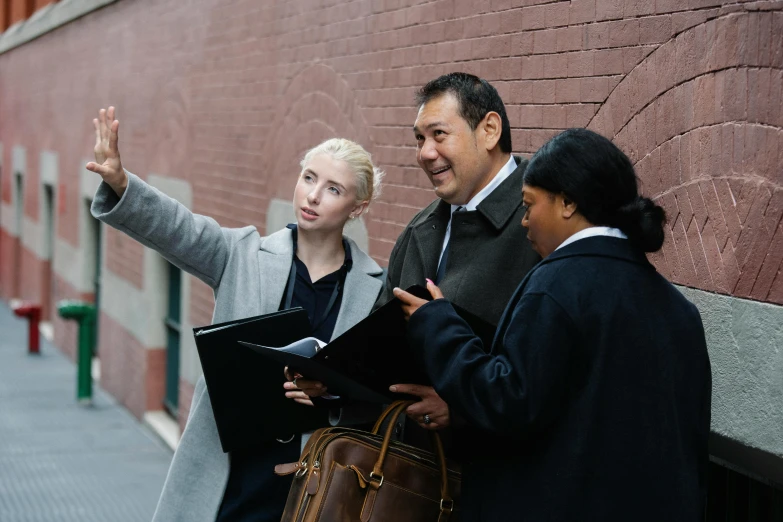 a group of people standing next to a brick wall, pointing, holding a briefcase, principal photography, street scene