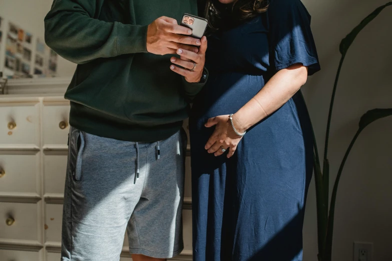 a man and a woman standing next to each other, trending on pexels, happening, pregnant, she is holding a smartphone, blue and grey, navy