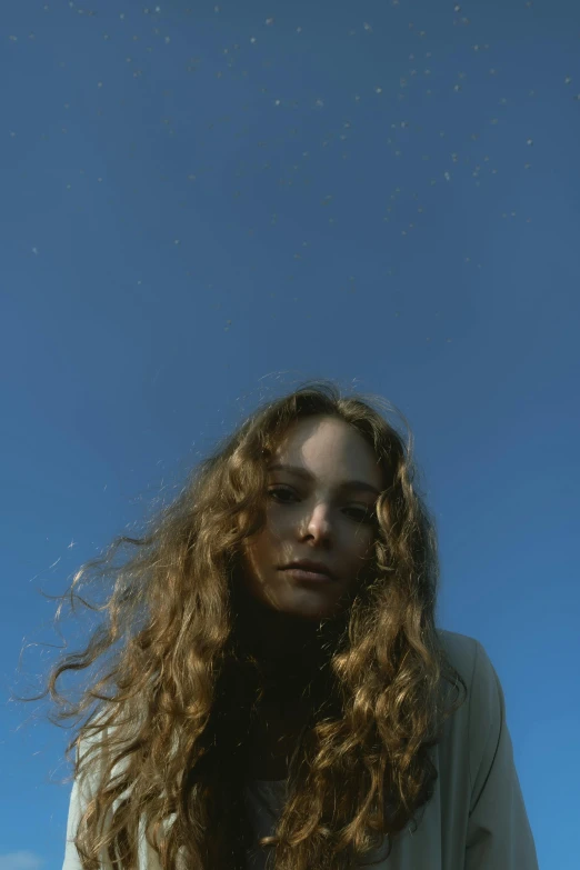 a woman standing in front of a blue sky, long curly brown hair, viewed from bellow, photo of young woman, ignant