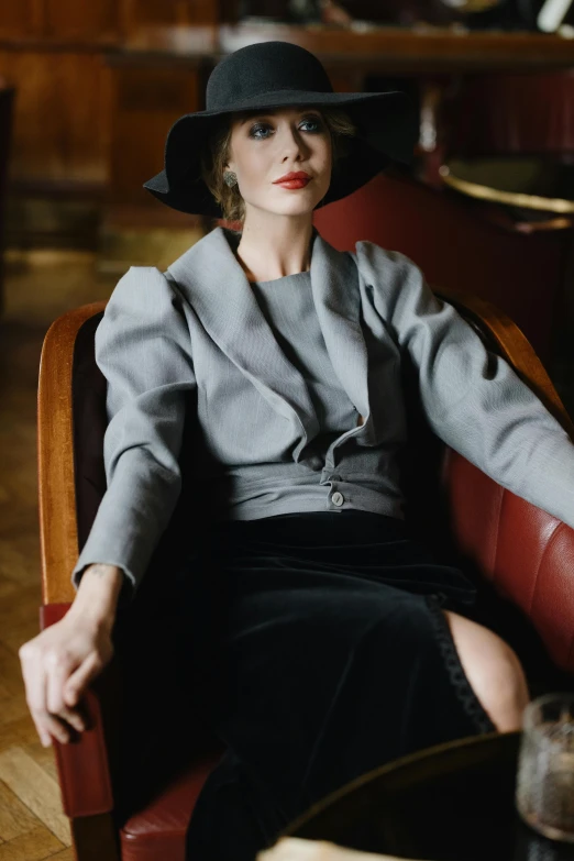 a woman sitting in a chair with a hat on, a portrait, inspired by August Sander, trending on pexels, grey jacket, in elegant decollete, in style of nan goldin, dressed in a gray