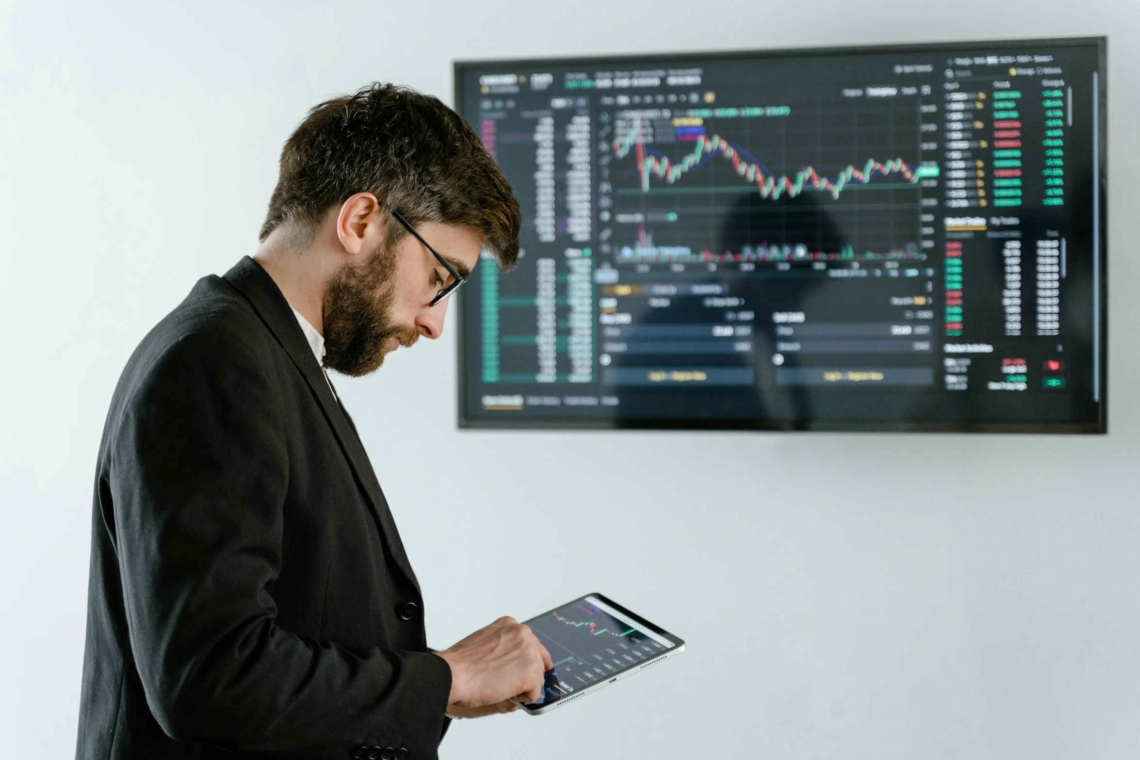 a man standing in front of a monitor holding a tablet, trending on pexels, analytical art, cryptocurrency, wearing causal black suits, australian, wearing a suit and glasses