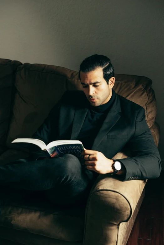 a man sitting on a couch reading a book, inspired by Nadim Karam, wearing causal black suits, christian saint, gal yosef, sebastian luca