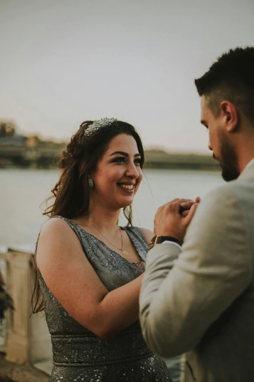 a man and a woman standing next to each other, pexels contest winner, wearing silver dress, river in the background, turning her head and smiling, half-turned lady in evening gown