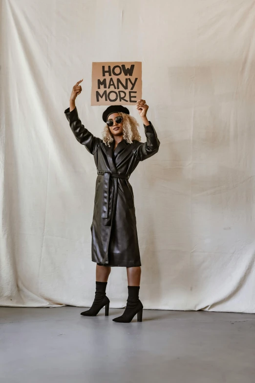 a woman standing in front of a white backdrop holding a sign, an album cover, by Emma Andijewska, trending on unsplash, maximalism, leather trench coat, performing, waving, nonbinary model