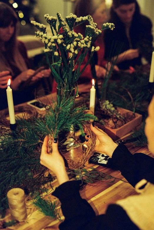 a group of people sitting around a table with candles, evergreen branches, in style of heikala, hand on table, hay