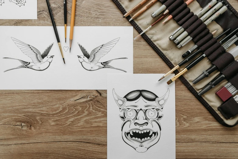 a couple of drawings sitting on top of a wooden table, an ink drawing, trending on pexels, monster design, heads of wooden of bird face, tattoo design, art set