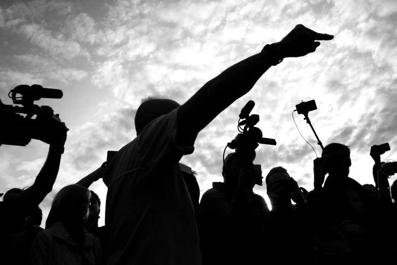 a black and white photo of a crowd of people with cameras, by Nadir Afonso, pexels, figuration libre, indistinct man with his hand up, microphone silluette, with pointing finger, skies behind