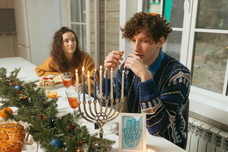 a man and woman sitting at a table with a menorah, by Julia Pishtar, pexels, finn wolfhard, wearing festive clothing, lifestyle, college