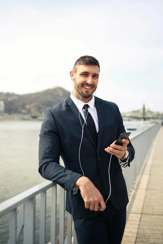 a man in a suit standing next to a body of water, an album cover, trending on pexels, happening, earbuds, on a bridge, flirting, hungarian