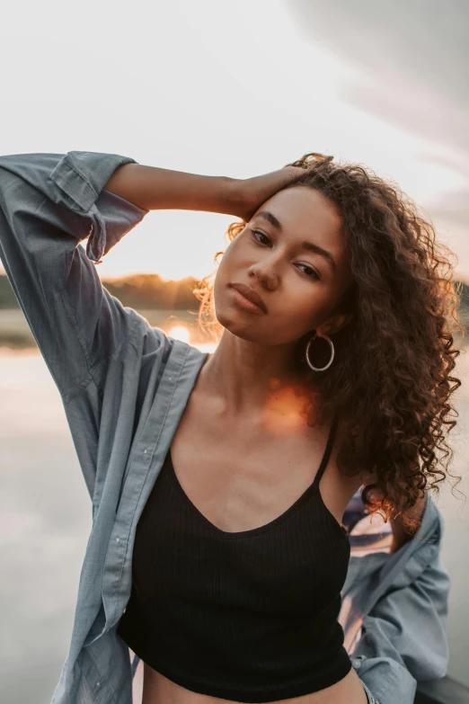 a woman standing next to a body of water, trending on pexels, renaissance, brown curly hair, sunset lighting, mixed race woman, on grey background