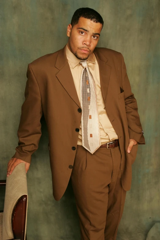 a man in a suit standing next to a chair, an album cover, inspired by Christopher Williams, harlem renaissance, wearing a worn out brown suit, photo from a promo shoot, circa 1 9 9 9, subject detail: wearing a suit