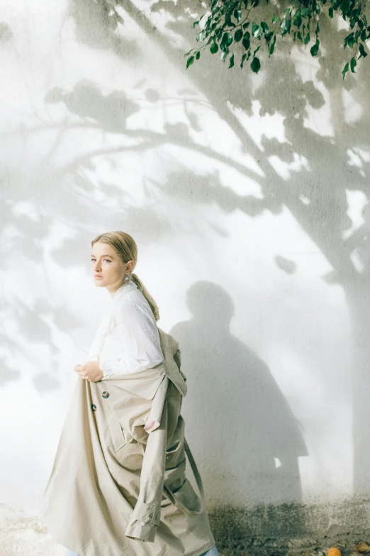 a woman standing in front of a white wall, by Caro Niederer, unsplash, visual art, under the soft shadow of a tree, light brown trenchcoat, sydney sweeney, sun-hyuk kim