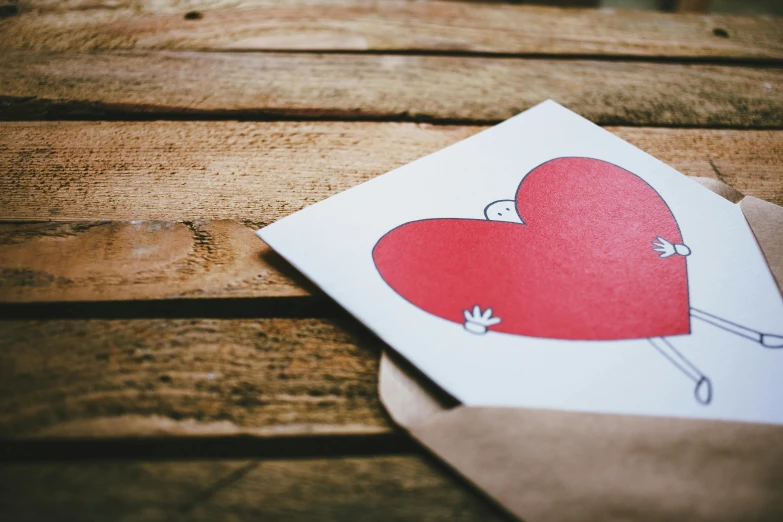 a piece of paper with a drawing of a heart on it, a cartoon, pexels contest winner, mail art, on a wooden table, focus on card, a red cape, trending photo