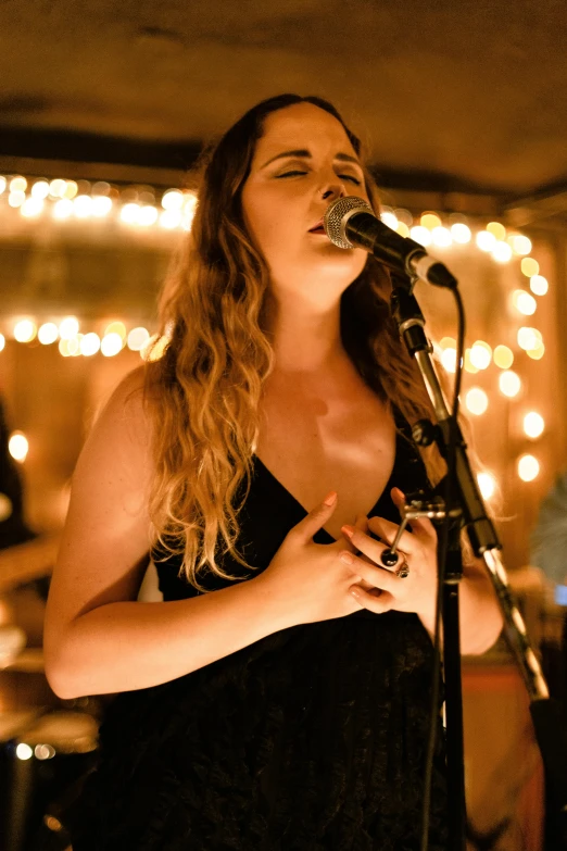 a woman in a black dress singing into a microphone, an album cover, pexels, happening, string lights, band playing, warm lighting inside, profile image