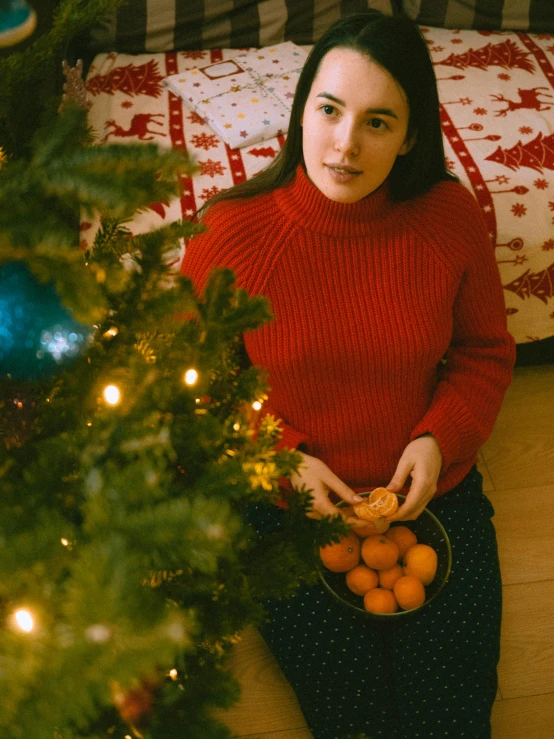 a woman sitting in front of a christmas tree holding oranges, by Julia Pishtar, pexels contest winner, red sweater and gray pants, gif, kramskoi 4 k, portrait sophie mudd