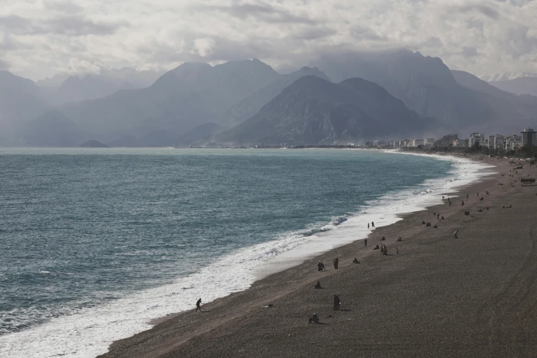 a group of people walking along a beach next to the ocean, inspired by Andreas Gursky, unsplash contest winner, baroque, mountains, turkey, grey, panoramic view