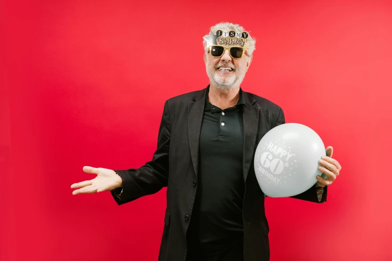 a man in a suit and sunglasses holding a white balloon, grey beard, celebrating a birthday, in red background, a silver haired mad